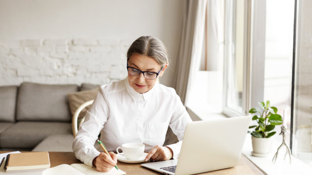 Picture of attractive mature woman business coach wearing white blouse and eyeglasses making notes in notebook while sitting at her cozy office, working on generic laptop computer and drinking coffee.