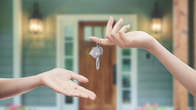 A hand passing house keys to another hand in front of a house.